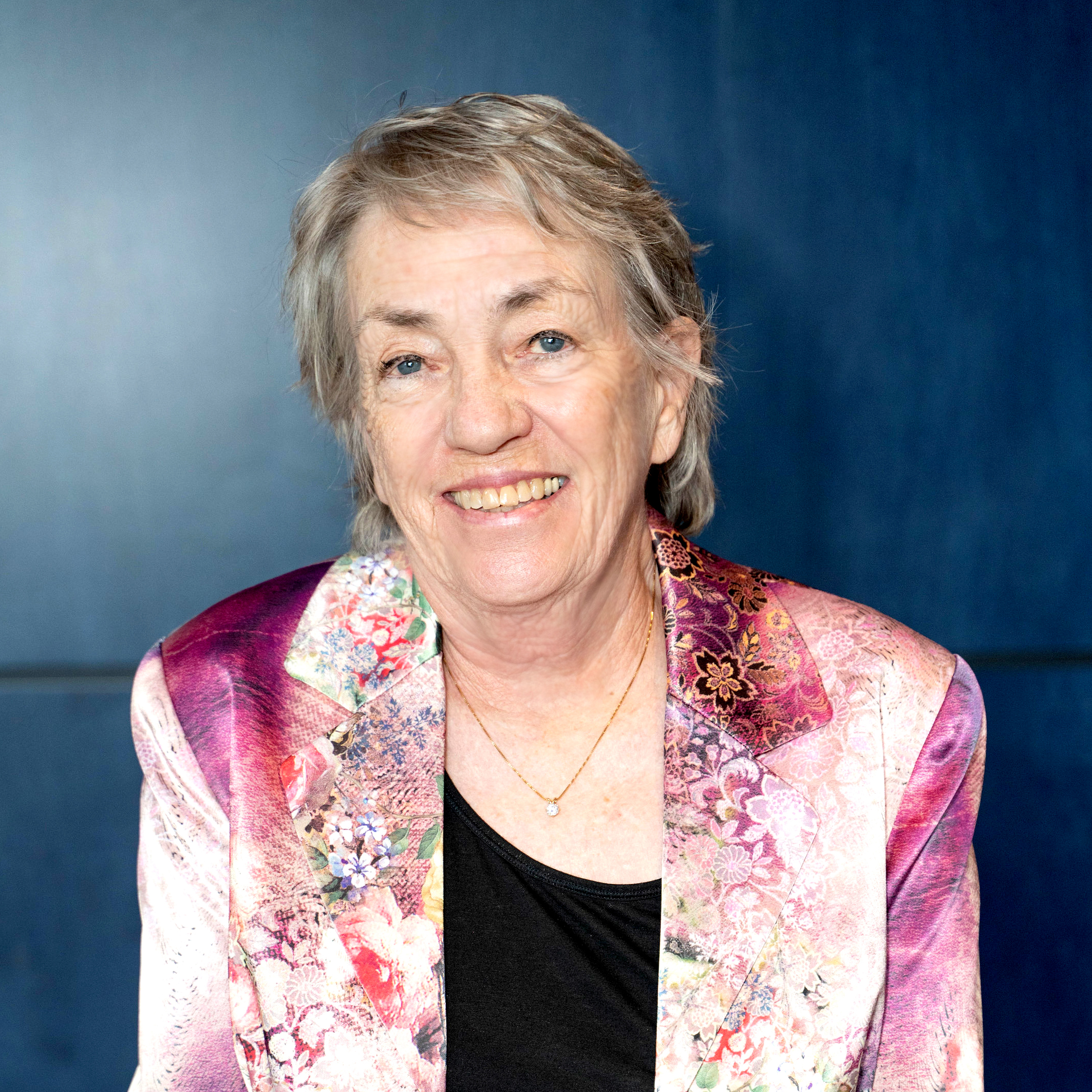 A headshot of Lucy Sanders smiling at the camera wearing a purple and white blazer over a black top in front of a gray-blue background