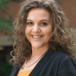 A headhshot of Kate Atchison wearing a dark blazer over an orange top, in front of a dark background