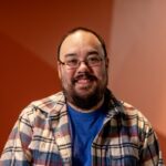 A headshot of Alan Jamieson wearing a plaid button down over a blue shirt, in front of an orange background