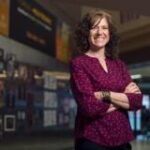 A photo of Dawn Laux wearing a crimson top with her arms crossed, standing in front of a blurred background