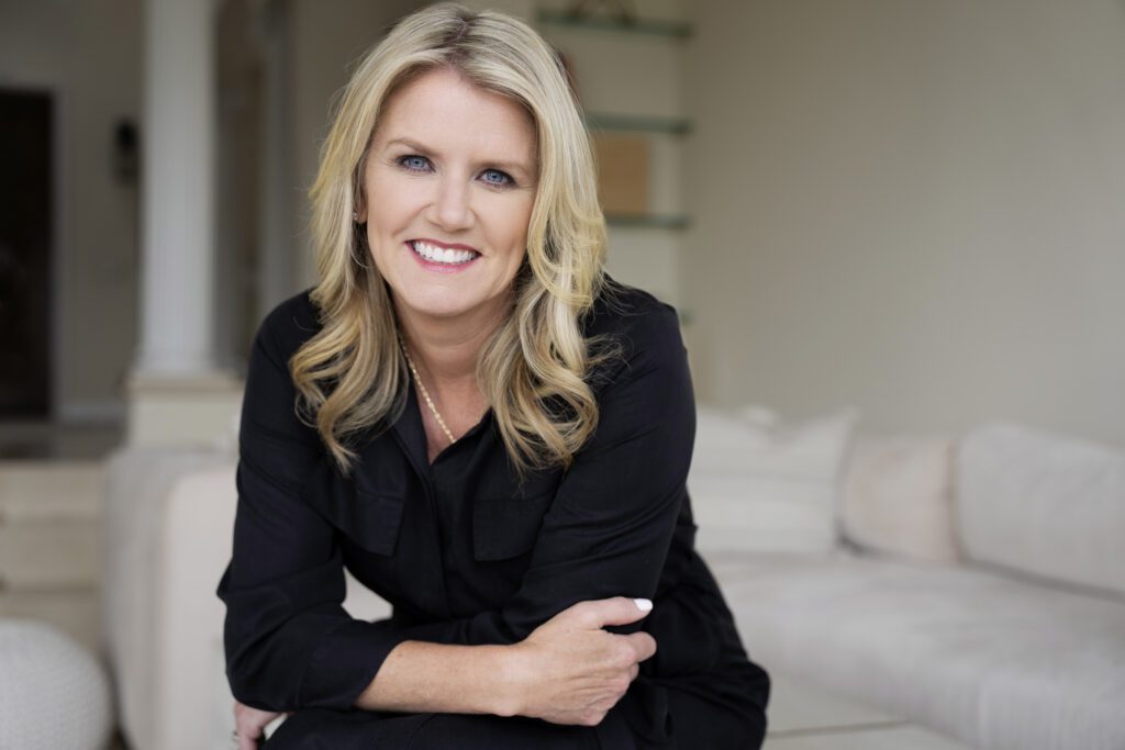 A headshot of Amy Coleman smiling at the camera. She's wearing a black shirt and standing in front of a tan couch