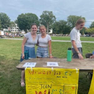 Color photo of Kaylie (R in blue shorts) and her colleague (L in a blue skirt) from Women in CS club fair standing in front of a yellow banner and brown table at a student organization fair