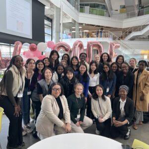 Smiling group photo of Kaylie — kneeling in front wearing a green sweater and khaki pants — with 25+ of diverse Women in Tech members at Harvard’s WeCode event