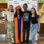 Color group photo of Neith — in a blue dress and orange graduation cap and gown — smiling with three other diverse student interns in front a “Amazon Future Engineer” banner