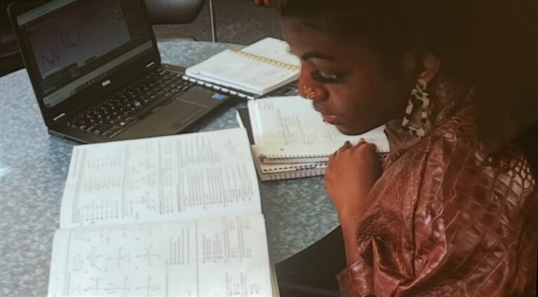 Vintage-like photo of Neith — in multi-colored earrings, a gold nose ring, and a brown jacket — studying pre-calculus at a desk with three notebooks and a computer