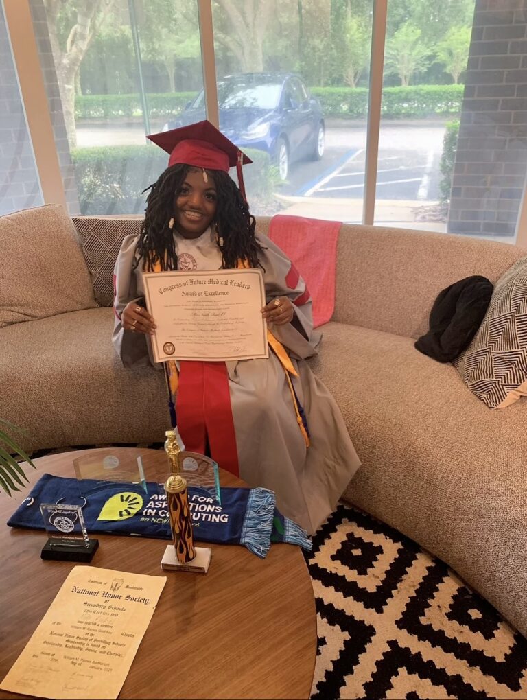 Photo of Neith smiling toward the viewer in a red and gray cap and gown receiving high school awards, including the NCWITAiC High School Award and the Congress of Future Medical Leaders certificate, with awards and saw on the brown table in front of her