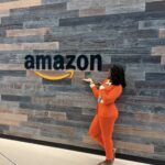 Vivid photo of Neith Reid-EL — while interning in Seattle, Washington — in an orange and yellow suit, smiling and gesturing toward the Amazon logo on the wall