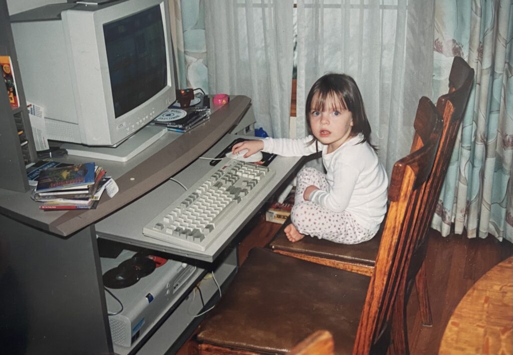 Color photo of a young Bryn — in a white pajama onesie — looking toward the viewer while playing at a desktop computer