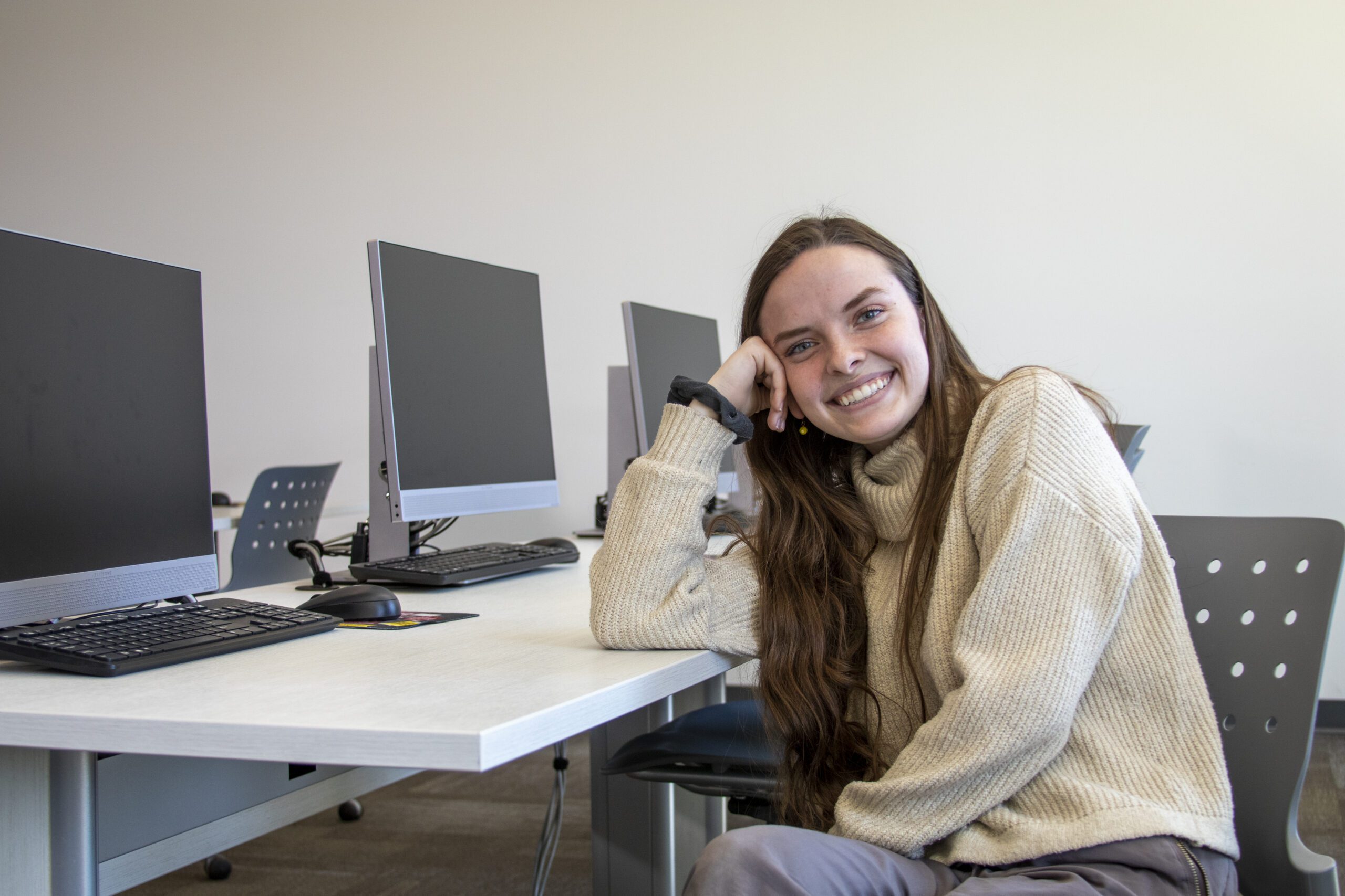 Color photo of Ph.D. candidate Bryn — with long brown hair and wearing a cream sweater with a black scrunchie on her wrist — smiling toward the viewer while sitting and leaning on a table with her head in her hand
