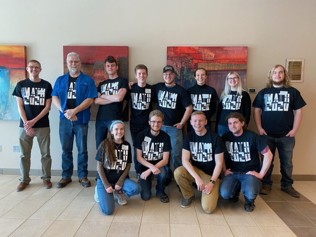 Color group photo of Bryn – first person kneeling on the left in a gray headband and blue jeans — smiling amongst 12 CS club members at Colorado Mesa University