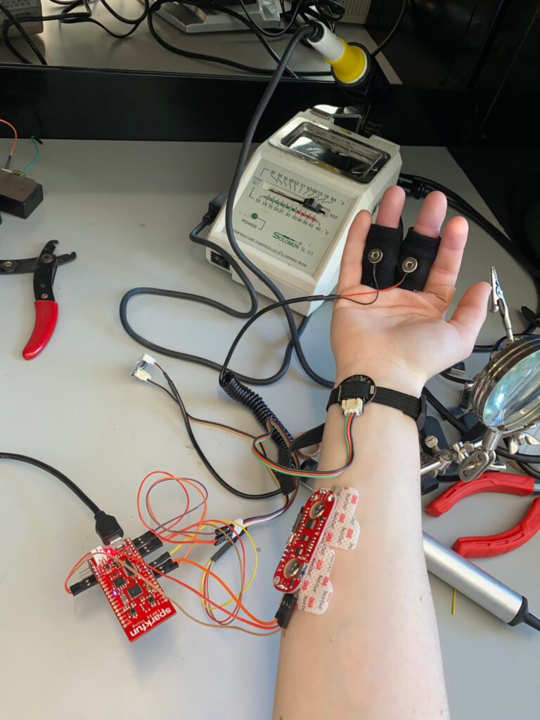 Color photo of Bryn holding out her arm while wearing her BPMBox — a pager-like homemade heart rate sensor — over a table with a white device, black and red cords, and two red clamp tools