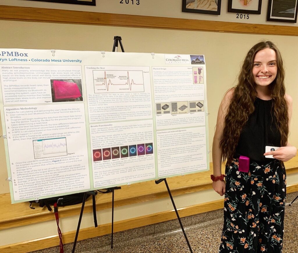 Color photo of Bryn — in a black blouse and floral skirt — smiling toward the viewer and pointing to a pink BMPBox pager on her hip while standing next to her presentation poster at an external NASA-sponsored research symposium