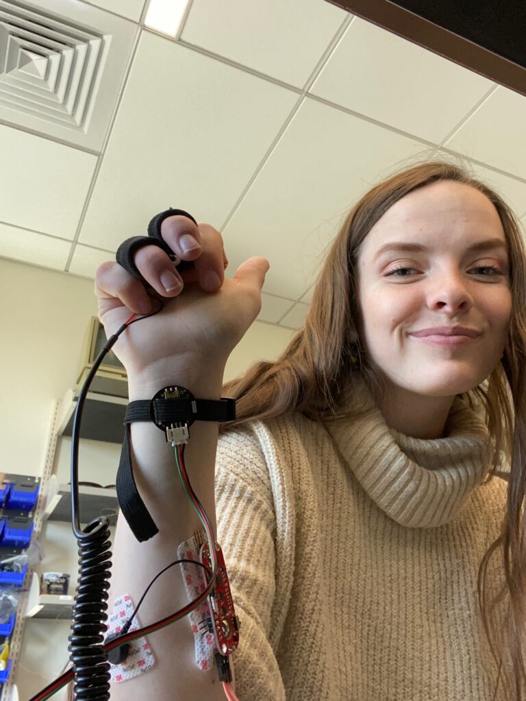 Bryn (in a turtleneck sweater) holding BPMBox — a pager-like homemade heart rate sensor that monitored pulse and provided bluetooth-based messages when a wearer of the device was undergoing abrupt heart rate changes