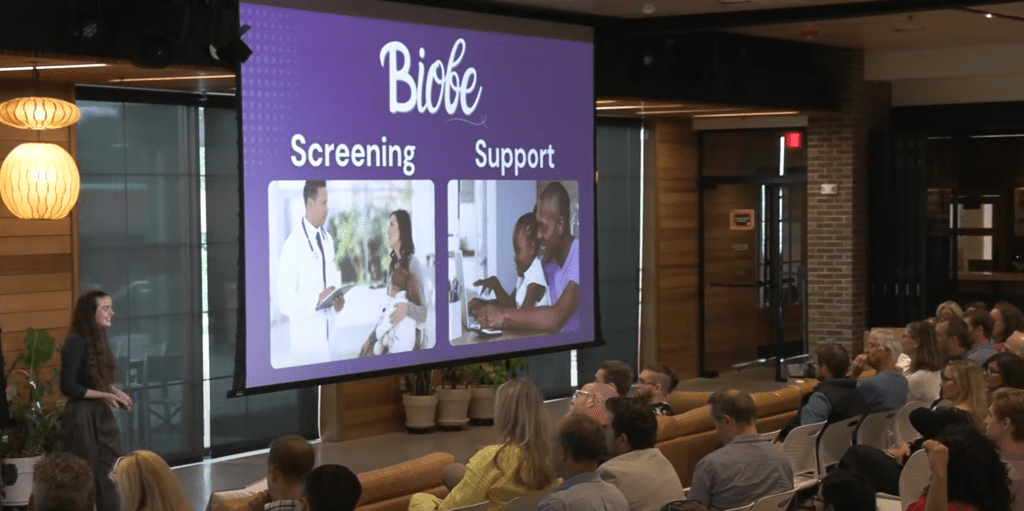 Behind-the-scenes snapshot of Bryn — in the left corner with long brown hair and trousers — presenting her start-up, Biobe, to a diverse crowd with a purple presentation screen with two images on display. Text reads: “Screening | Support”