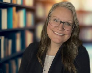 A headshot of Jenny Frank smiling at the camera in front of bookshelves